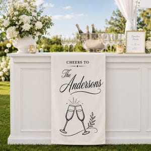 May include: A white fabric table runner featuring black printed text that reads "CHEERS TO The Andersons" above a graphic illustration of two clinking champagne flutes with a small leafy branch detail at the bottom of the design.