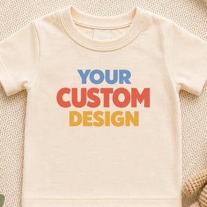 May include: A cream-coloured short-sleeve t-shirt featuring the text "YOUR CUSTOM DESIGN" printed in blue, red, and yellow block letters on the front. The shirt has a crew neckline and a straight hem, presented with a small crocheted teddy bear.