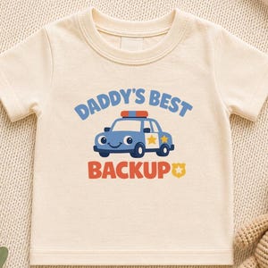 May include: A cream-coloured short-sleeve t-shirt featuring a graphic of a blue police car with a smiling face. The text above the car reads DADDY'S BEST and the text below reads BACKUP with a small yellow star icon next to the word backup.