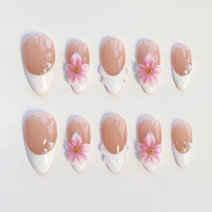 Può includere: Un set di dieci unghie finte con base color carne e punte bianche alla francese. Alcune unghie sono decorate con fiori di ciliegio rosa tridimensionali con centri dorati, mentre altre presentano piccole perle bianche e perline dorate.