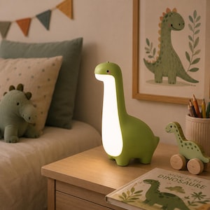 May include: A light green dinosaur-shaped lamp with a smooth, rounded body and a glowing white front panel. The lamp stands on a wooden surface, emitting a soft, warm light from its chest area, featuring a simple, minimalist design with a small pink cheek.