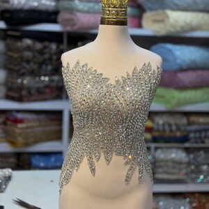 May include: A sleeveless bodice applique featuring an intricate arrangement of clear, faceted crystals and silver-toned beads on a sheer nude mesh base. The design showcases a leaf-like pattern that extends downward, creating a pointed hemline.