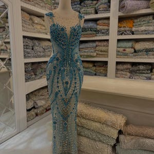 Könnte beinhalten: Ein bodenlanges ärmelloses Kleid mit einer transparenten, hautfarbenen Basis, die mit aufwendigen blauen Perlen, Pailletten und Kristallen verziert ist. Das Kleid hat eine figurbetonte Silhouette, einen Rundhalsausschnitt und florale Muster.