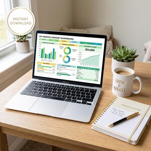 May include: A silver laptop displaying a My Monthly Budget Dashboard with charts and financial data. Beside it sits a white mug labeled Brew & Budget, a spiral-bound notebook with handwritten July Goals, and a small potted succulent plant.