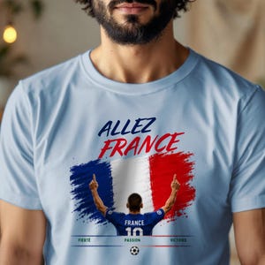 May include: A light blue t-shirt featuring a graphic of the French flag in a brushstroke style. Above the flag, the text reads "ALLEZ FRANCE". Below, a soccer player in a blue jersey with "FRANCE 10" stands with arms raised, with "FIERT&Eacute;", "PASSION", and "VICTOIRE" printed below.