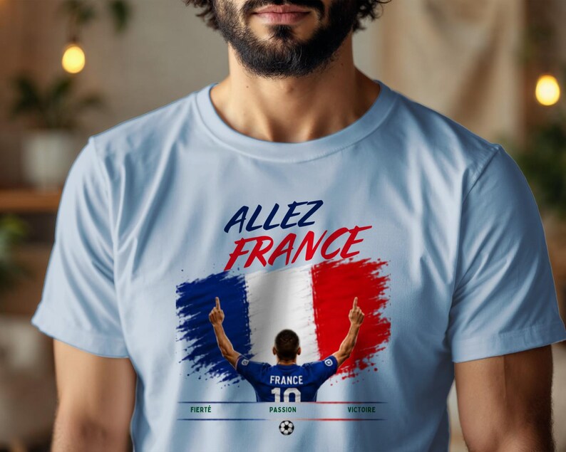 May include: A light blue t-shirt featuring a graphic of the French flag in a brushstroke style. Above the flag, the text reads "ALLEZ FRANCE". Below, a soccer player in a blue jersey with "FRANCE 10" stands with arms raised, with "FIERT&Eacute;", "PASSION", and "VICTOIRE" printed below.