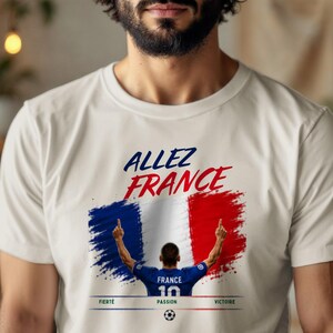 May include: A cream-colored t-shirt featuring a graphic print with the text "ALLEZ FRANCE" in blue and red. Below, a stylized French flag background sits behind a soccer player figure, with the words "FIERT&Eacute;", "PASSION", and "VICTOIRE" below.