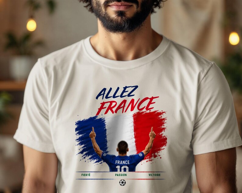 May include: A cream-colored t-shirt featuring a graphic print with the text "ALLEZ FRANCE" in blue and red. Below, a stylized French flag background sits behind a soccer player figure, with the words "FIERT&Eacute;", "PASSION", and "VICTOIRE" below.