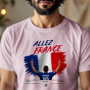 May include: A light pink t-shirt featuring the text "ALLEZ FRANCE" in blue and red. Below, a graphic shows a soccer player in a blue jersey with the number 10, standing before a French flag design. The words "FIERT&Eacute;", "PASSION", and "VICTOIRE" appear below.
