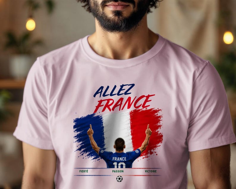 May include: A light pink t-shirt featuring the text "ALLEZ FRANCE" in blue and red. Below, a graphic shows a soccer player in a blue jersey with the number 10, standing before a French flag design. The words "FIERT&Eacute;", "PASSION", and "VICTOIRE" appear below.