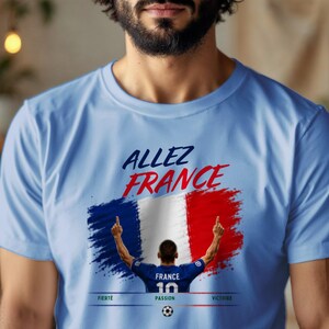 May include: A light blue t-shirt featuring a graphic of the French flag with the text "ALLEZ FRANCE" in blue and red. Below the flag is an illustration of a soccer player wearing a blue jersey with the number 10, and the words "FIERT&Eacute;", "PASSION", and "VICTOIRE".