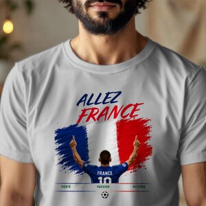 May include: A light gray t-shirt featuring a graphic of the French flag in a brushstroke style. Above the flag, the text reads ALLEZ FRANCE. Below, a soccer player in a blue jersey with the number 10 is shown, with the words FIERT&Eacute;, PASSION, VICTOIRE.