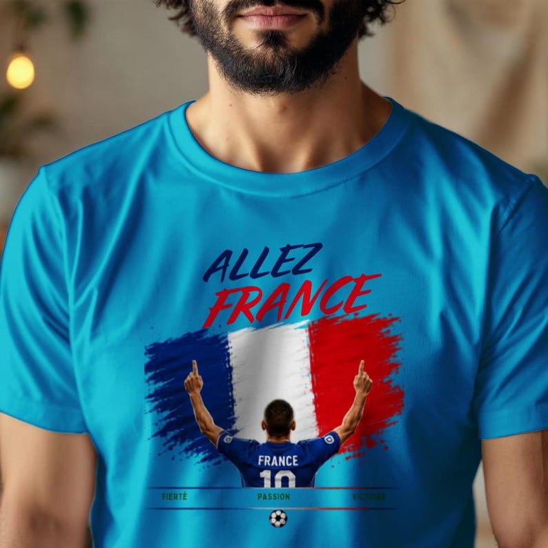 May include: A bright blue t-shirt featuring the text "ALLEZ FRANCE" in dark blue and red. Below, a graphic shows a soccer player in a blue jersey with "FRANCE 10" on the back, standing before a French flag. The words "FIERT&Eacute;", "PASSION", and "VICTOIRE" appear below.