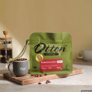 May include: A green pouch of Otten Coffee Robusta Aceh with a 200g net weight label, accompanied by a grey mug filled with coffee beans. The pouch features a yellow QR code sticker, text detailing the region and altitude, and a green rope handle.