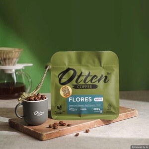 May include: An olive green coffee bag labelled Otten Coffee Flores with a 200g weight, featuring a QR code sticker, next to a small grey mug filled with roasted coffee beans. The bag is connected to the mug by a patterned cord.