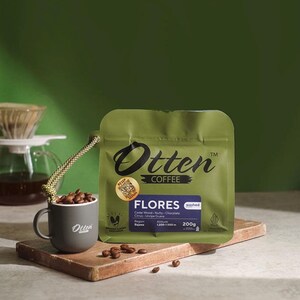May include: An olive green coffee bag labelled Otten Coffee Flores with a net weight of 200g, featuring a QR code sticker. Beside it sits a small grey mug filled with roasted coffee beans, connected to the bag by a braided cord.