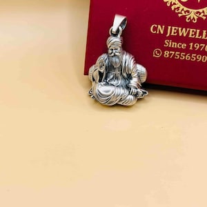 May include: A silver pendant depicting a seated figure with a long beard and turban. The pendant is detailed with intricate carvings. The background includes a red box with the text "CN JEWELL" and a phone number.
