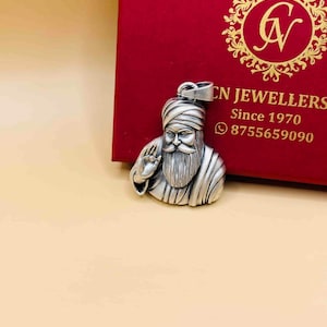 Può includere: Un ciondolo in metallo color argento raffigurante una figura barbuta con turbante, posizionato davanti a una scatola rossa con testo dorato CN JEWELLERS Since 1970 e un'icona del telefono seguita dal numero 8755659090.
