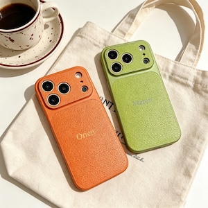 Op de afbeelding: Twee getextureerde telefoonhoesjes, één oranje en één groen, met een uitsparing voor de camera en de namen "Orien" en "Margo" op de achterkant gedrukt. Het oranje hoesje staat links en het groene hoesje staat rechts.