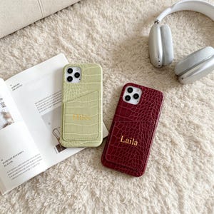 Può includere: Due custodie rettangolari per smartphone con una texture a motivo pelle di coccodrillo. Una custodia è verde chiaro con il nome Miles impresso in oro, l'altra è rosso scuro con il nome Laila impresso in oro. Ogni custodia ha uno scomparto per carte.