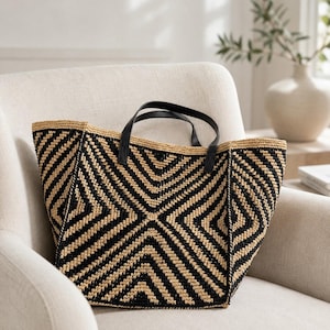 May include: A woven tote bag featuring a geometric diamond pattern in alternating black and natural tan straw-like material. The bag includes two black leather-like handles attached to the top edge, with a structured shape and a wide opening.