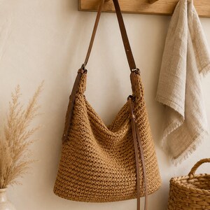 May include: A tan crochet hobo bag featuring a textured, woven pattern and a long, adjustable brown leather shoulder strap. The bag includes a thin leather drawstring closure with two dangling leather cords hanging from the side of the opening.
