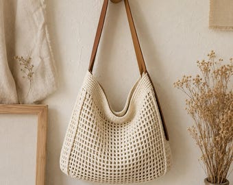 May include: A cream-colored crochet hobo bag featuring a square grid pattern throughout the body. The bag includes a single, smooth brown leather shoulder strap attached to the top corners, creating a slouchy, open-top silhouette for carrying.