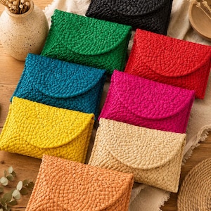 Puede incluir: Una colección de ocho bolsos de mano rectangulares tipo sobre hechos de material trenzado en una variedad de colores vibrantes que incluyen negro, verde, rojo, azul, magenta, amarillo, beige y naranja, cada uno con un cierre de solapa triangular.