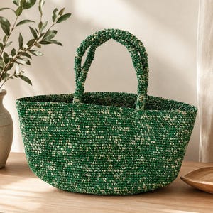 May include: A woven tote bag featuring a textured, variegated pattern of deep forest green and off-white fibres. The bag has a rounded, open-top silhouette and two sturdy, matching woven handles attached to the upper rim for carrying.