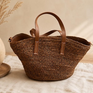 May include: A dark brown woven straw basket bag featuring a wide, open top and two smooth, tan-coloured leather handles attached with white stitching. The bag has a rounded, structured base and gold-toned metal hardware rings on the sides.