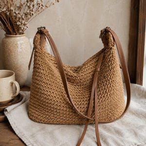 May include: A tan crochet shoulder bag featuring a textured, woven knit pattern and a smooth brown leather strap with an adjustable metal buckle. The bag has a soft, slouchy silhouette and includes long, thin leather cord accents hanging down.