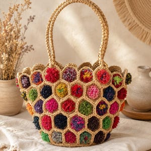 May include: A woven straw tote bag featuring a hexagonal honeycomb pattern. Each hexagon contains a colourful yarn pom-pom in shades of red, pink, yellow, green, blue, and purple. The bag has two braided straw handles and a scalloped top edge.