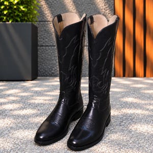 May include: A pair of black knee-high boots featuring a pointed toe, a low block heel, and intricate tonal stitching patterns on the shaft and vamp. The boots have a classic pull-on design with a V-shaped topline and a light-coloured interior lining.