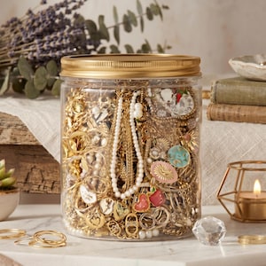 May include: A clear plastic cylindrical jar with a gold-colored metal lid, filled with a collection of gold-toned jewelry including chains, rings, pearl-like beaded strands, and various colorful enamel charms featuring heart and floral motifs.