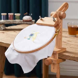 Puede incluir: Un soporte de madera clara para bastidor de bordado con mecanismo de abrazadera ajustable, que sostiene un bastidor circular de madera con tela blanca que presenta un pequeño diseño floral parcialmente bordado en hilo amarillo, verde y morado.
