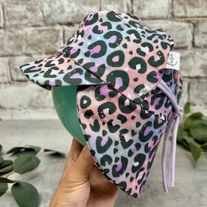Könnte beinhalten: Ein Sonnenhut mit einem pastellfarbenen Farbverlauf in Rosa, Lila und Blau sowie einem schwarzen Leopardenmuster. Der Hut verfügt über eine feste Krempe, einen Nackenschutz und helllila Stoffbänder. Ein kleines weißes Etikett trägt die Aufschrift Jellyfish.