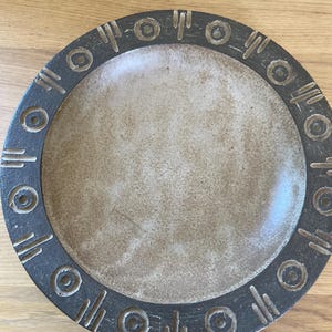 May include: A round, ceramic plate with a textured, beige interior and a dark gray border. The border features a repeating pattern of circles and lines. The plate is likely used for serving food or as a decorative piece.
