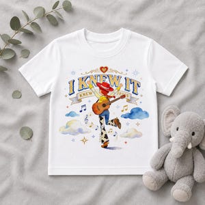May include: A white short-sleeved t-shirt featuring a graphic of a cowgirl character playing an acoustic guitar. The design includes the text I KNEW IT, musical notes, clouds, and stars in blue, yellow, and red colors. The character wears a red hat.