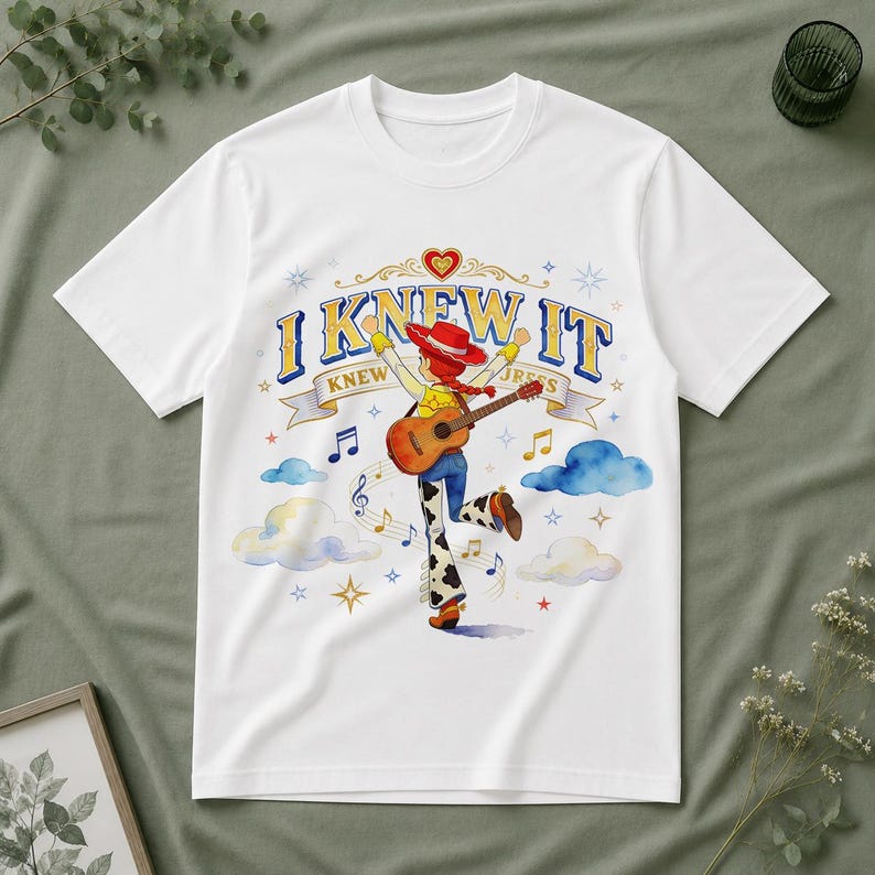 May include: A white short-sleeve t-shirt featuring a graphic of a cowgirl character with red hair playing an acoustic guitar. The text I KNEW IT is printed above the character, surrounded by blue clouds, musical notes, and yellow stars.