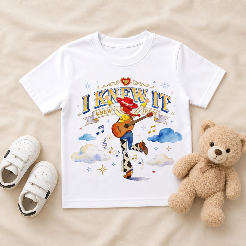 May include: A white short-sleeved t-shirt featuring a graphic of a cowgirl character with a red hat and guitar, surrounded by clouds, musical notes, and stars. The text "I KNEW IT" is printed in blue and yellow above the character on the shirt.