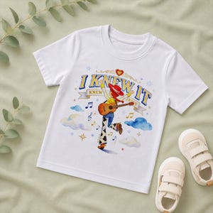 May include: A white short-sleeved t-shirt featuring a graphic of a cowgirl character playing an acoustic guitar. The text "I KNEW IT" is printed above the character, surrounded by blue clouds, musical notes, and small star-shaped accents.