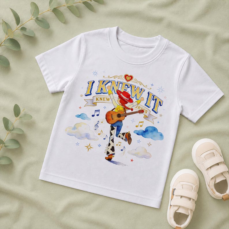 May include: A white short-sleeved t-shirt featuring a graphic of a cowgirl character playing an acoustic guitar. The text "I KNEW IT" is printed above the character, surrounded by blue clouds, musical notes, and small star-shaped accents.