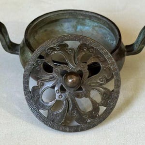 May include: A dark bronze incense burner with two side handles and a removable circular lid. The lid features an intricate openwork floral pattern surrounded by a geometric border, while the bowl has a smooth, weathered patina finish.