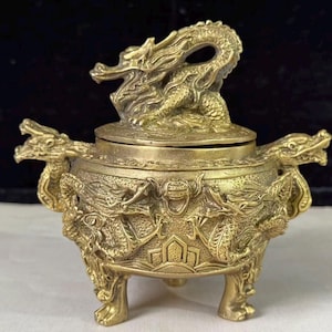 May include: A gold-colored metal incense burner featuring a lid topped with a sculpted dragon figure. The round vessel is supported by three legs and decorated with relief carvings of dragons on the sides and two dragon-shaped handles.