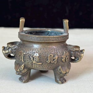 May include: A bronze-toned metal incense burner featuring a textured surface, two upright handles, and three legs shaped like animal heads. The body is decorated with four raised Chinese characters and a geometric border pattern around the rim.