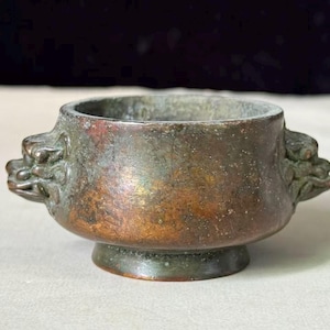 May include: A small, round bronze vessel with a weathered, dark patina and a metallic copper-toned finish. The bowl features two sculpted animal-head handles on opposite sides and rests on a circular pedestal base with a wide, open top rim.