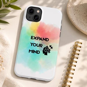 Puede incluir: Una funda de smartphone blanca con un fondo estilo acuarela con salpicaduras mezcladas de amarillo, rosa y turquesa. El texto "EXPAND YOUR MIND" está impreso en negro, acompañado por una silueta negra de una cabeza humana con piezas de rompecabezas.