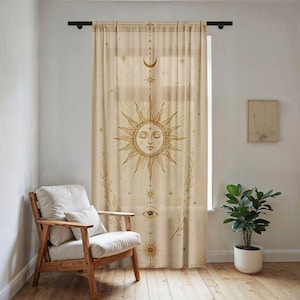 May include: A sheer beige curtain hanging from a black rod, featuring a gold-toned print of a central sun face with closed eyes, surrounded by rays, stars, a crescent moon, and an eye symbol, with vertical botanical accents along the sides.