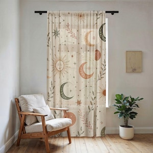 May include: A sheer window curtain featuring a repeating pattern of celestial motifs including suns, moons, and stars in shades of orange, green, and brown on a cream background, suspended from a black rod over a wooden floor.