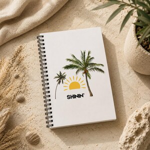 Puede incluir: Cuaderno blanco con espiral con un diseño de palmera y sol. La palabra "SHININ'" está impresa debajo del sol. El cuaderno está sobre una superficie arenosa, con una tela beige y una planta en maceta en el fondo.